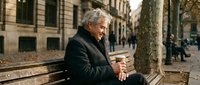 Senior Caucasian businessman in elegant coat sitting on urban bench using smartphone in historic European city street during autumn day.