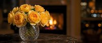 Golden yellow roses in crystal vase on wooden table with warm fireplace background creating cozy romantic home interior atmosphere for decoration.
