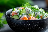 Fresh salmon salad with cucumber, dill and herbs in black speckled bowl. Healthy seafood dish with mixed greens for restaurant menu or cooking blog.