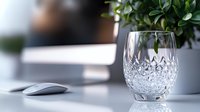 Crystal whiskey glass on modern desk with computer mouse and green plant, elegant home office setup in soft natural lighting. Professional workspace decor.