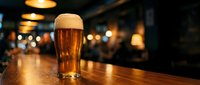 Fresh pint of golden beer with creamy foam head on wooden bar counter in cozy pub atmosphere with warm ambient lighting and blurred background.