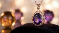 Elegant amethyst oval pendant with diamond halo on silver chain, displayed on black velvet with blurred gemstone bokeh background. Luxury jewelry photography.