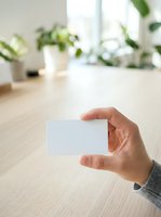 Close-up of a hand holding a blank white business card in a bright office workspace with potted plants and natural light from windows.