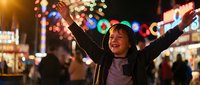 Young Asian woman celebrating with raised arms at vibrant night market with colorful bokeh lights and festive atmosphere for holiday marketing campaigns.
