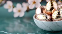 Golden speckled Easter eggs in marble bowl with soft focus white spring blossoms against teal background, creating festive holiday atmosphere.