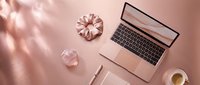 Flat lay of a rose gold laptop, silk scrunchie, crystal, notebook, pen and coffee cup on a pink desk, feminine workspace top view.