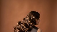 Young woman with long, voluminous wavy brown hair styled in loose waves, shown in profile against warm brown background. Suitable for hair care, beauty, and wellness marketing.