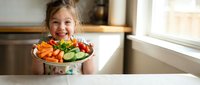 Happy young girl holding colorful plate of fresh vegetables in bright kitchen promoting healthy eating habits for children and nutritious lifestyle.