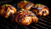 Juicy grilled chicken thighs wrapped in crispy bacon, glazed with sweet and savory sauce, garnished with fresh herbs on dark barbecue grill grates.