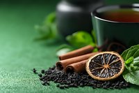 Aromatic black tea composition with dried citrus, cinnamon sticks, fresh mint leaves and loose tea on green textured background with copy space. Cozy tea time setting.
