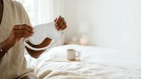 Woman with medium brown skin tone holding empty bowl and wooden spoon while sitting on bed in bright bedroom, expressing hunger or disappointment about empty meal.