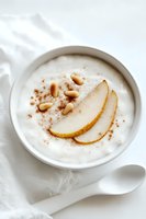 Creamy white yogurt bowl topped with fresh pear slices, pine nuts and cinnamon powder on white background, served with ceramic spoon. Healthy breakfast concept.