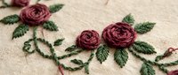 Hand embroidered dark red roses with green leaves on natural linen fabric, showcasing intricate needlework and floral stitch patterns for craft inspiration.