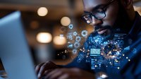 African American male professional working on laptop with glowing digital interface overlay showing technology connections and data visualization elements in dark office setting.