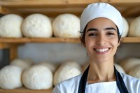 Young female baker in chef uniform smiling at bakery with fresh bread dough balls on wooden shelves in background, representing small business and artisanal baking.