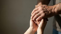 Elderly wrinkled hands gently holding young smooth hands showing intergenerational connection, care and support between generations in warm lighting.