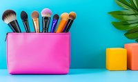 Colorful makeup brushes in bright pink cosmetic bag against turquoise background with tropical leaf and geometric shapes, modern beauty concept.