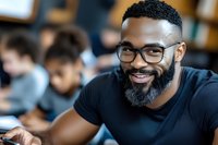 African American male professional wearing glasses and black t-shirt smiles warmly during workplace meeting or educational setting. Blurred coworkers in background.