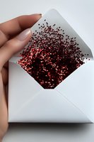 Red glitter hearts spilling out of white envelope held by feminine hand with neutral manicure against light background, creating magical romantic effect.