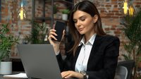 Young businesswoman in formal suit working on laptop and checking smartphone at modern loft office with brick wall and decorative lighting. Professional workspace concept.