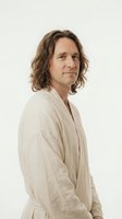 Middle-aged Caucasian man with shoulder-length wavy brown hair wearing beige linen robe against neutral background for wellness and spa concepts.