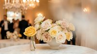 Elegant wedding centerpiece with cream and yellow roses in white vase on reception table with chandelier and guests in blurred background.