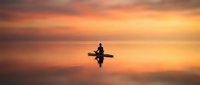 Silhouette of person kayaking on calm water during golden sunset with dramatic orange sky reflecting on lake surface creating peaceful outdoor recreation scene.
