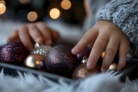 Child's hands touching glittering Christmas ornaments in purple and rose gold colors against soft bokeh lights background, creating cozy holiday atmosphere.