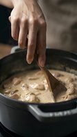 Close-up of hand stirring creamy mushroom sauce in cast iron skillet with wooden spoon for cooking and culinary preparation.