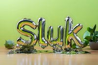 Metallic silver balloon letters spelling "Sluk" on wooden surface with green background and small potted plant, modern typography concept.