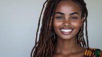 Young African woman with long braided hair and gold hoop earrings smiling at camera against neutral background for portrait, headshot, beauty, and professional use.
