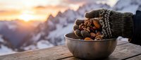 Hands in knitted gloves holding bowl of nuts against snowy mountain landscape at sunrise, winter outdoor activity and seasonal warmth concept.