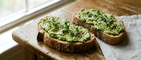 Two slices of rustic sourdough bread topped with creamy avocado spread, sesame seeds, and fresh microgreens on a wooden board.