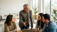 Mature male professional with gray hair, Caucasian ethnicity, leading business meeting with diverse team of younger colleagues in modern office environment with natural lighting and plants.