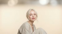 Young blonde Caucasian woman in beige linen shirt sitting relaxed with soft natural lighting and neutral background for lifestyle photography.