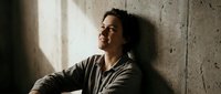 Young Hispanic man in casual gray sweater leaning against concrete wall with natural window light creating dramatic shadows on face in contemplative pose.