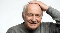 Senior man with light skin tone, bald head, wearing gray sweater, hand touching head in thoughtful pose, smiling at camera against white background for portrait use.