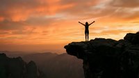Person celebrating success on mountain cliff edge during golden sunset with arms raised in victory pose overlooking scenic valley landscape.