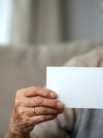 Senior adult male with wrinkled skin holding blank white card, wearing gold ring. Closeup of aged hand displaying empty paper for messaging or branding purposes.
