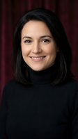 Confident middle-aged Hispanic woman in black turtleneck smiling against dark red curtain backdrop, professional portrait for business use.