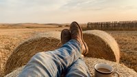 Person relaxing on hay bale in golden wheat field during sunset with coffee cup, peaceful rural countryside scene for agriculture and lifestyle content.