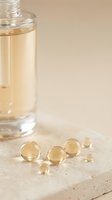 Glass jar filled with pale golden liquid and scattered translucent gel capsules on neutral stone surface. Suitable for wellness, supplement, and skincare product marketing.