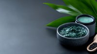 Green clay mask in black ceramic bowl with tropical leaf on dark background. Natural skincare and spa treatment for facial cleansing and detox procedures.