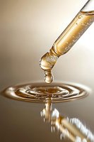Golden liquid drop falling into rippling surface creating concentric circles and splash reflection in warm tones against neutral background.