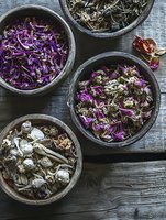 Dried purple and white flower petals in rustic ceramic bowls on wooden surface, moody natural lighting creates atmospheric composition for wellness and spa concepts.