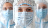 Healthcare worker in protective equipment with face shield and surgical mask during pandemic, focused expression with colleagues in background.