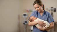 Female nurse in blue scrubs holding newborn baby in hospital room with medical equipment in background for healthcare and maternity concepts.