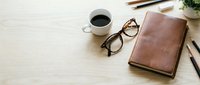 Cozy workspace flat lay with leather notebook, coffee cup, glasses, and pencils on a light wooden desk with copy space.