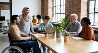 Diverse team collaborating in modern office with brick walls. Includes wheelchair user, hijab-wearing woman, and mixed ethnicities working together professionally.