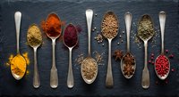 Colorful spices and seasonings arranged in vintage metal spoons on dark slate background for culinary cooking and food preparation projects.